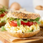 A close-up of a delicious Veggie Breakfast Sandwich on an everything bagel, featuring scrambled eggs, fresh tomatoes, and arugula on a wooden board.