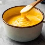 A wooden spoon stirs a rich, creamy yellow Velveeta Cheese Sauce in a white bowl on a grey background.