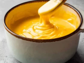 A wooden spoon stirs a rich, creamy yellow Velveeta Cheese Sauce in a white bowl on a grey background.