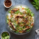 An overhead view of a large clear bowl filled with Vietnamese Noodle Salad with Grilled Chicken, garnished with mint and peanuts.