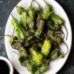 A white plate piled high with perfectly blistered green Air Fryer Shishito Peppers, ready to be enjoyed with a dipping sauce.
