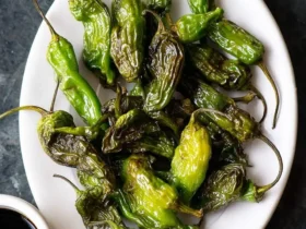 A white plate piled high with perfectly blistered green Air Fryer Shishito Peppers, ready to be enjoyed with a dipping sauce.