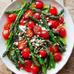 A vibrant Asparagus Tomato Salad on a white plate, featuring tender green asparagus, red cherry tomatoes, feta, and pine nuts.
