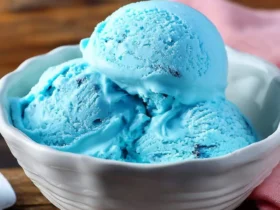 A white bowl holds three scoops of vibrant blue Blue Moon ice cream with dark flecks, set on a rustic wooden table.