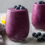 A vibrant purple Blueberry Avocado Smoothie in a glass, topped with fresh blueberries, on a marble counter with a lemon slice.