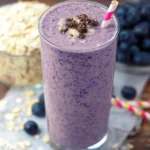 A tall glass of purple blueberry oatmeal smoothie topped with oats and granola, with a striped straw.