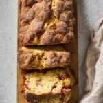A delicious loaf of Buttermilk Rhubarb Bread, sliced and showing its tender crumb and pink rhubarb pieces, on a wooden board.