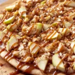 A close-up of a delicious Caramel Apple Dessert Pizza topped with green apple slices, chopped nuts, and rich caramel drizzle.