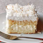 A delightful slice of Coconut Cream Cake topped with fluffy white frosting and toasted shredded coconut on a white plate with a fork.
