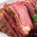 Close-up of perfectly sliced Corned Beef Brisket with a flavorful crust and a sprig of parsley on a white plate.