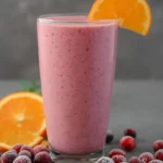 A tall glass of pink cranberry orange smoothie garnished with an orange slice, surrounded by frosted cranberries and orange halves.