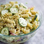 A clear glass bowl filled with creamy Cucumber Pasta Salad, featuring spiral pasta, fresh cucumber slices, and red onion.