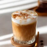 Iced Dirty Chai Latte in a glass with foamed milk, cinnamon sprinkles, on a coaster with coffee beans and a cinnamon stick.