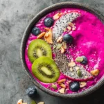 A vibrant pink Dragon Fruit Smoothie Bowl topped with kiwi, blueberries, dragon fruit slices, chia seeds, almonds, and pumpkin seeds on a dark grey background.
