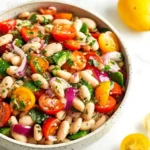 A vibrant Greek Bean Salad in a speckled bowl featuring white beans, halved cherry tomatoes, red onion, green bell pepper, and fresh herbs.