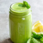 A vibrant green keto smoothie in a mason jar, garnished with fresh mint, alongside lemon, cucumber, and spinach on a grey surface.