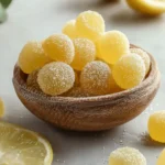A wooden bowl overflowing with bright yellow, sugar-coated lemon drops, with fresh lemon slices nearby on a grey surface.