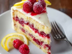A tempting slice of lemon raspberry cake on a white plate, garnished with fresh raspberries and lemon slices.
