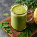 A vibrant green, frothy Moringa smoothie in a glass mug, placed on a wooden board with fresh moringa leaves and a banana.