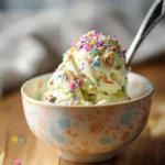 A bowl of creamy Ninja Creami Birthday Cake Ice Cream, topped with colorful sprinkles, with a spoon resting inside.