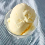 Three scoops of creamy light yellow Ninja Creami Pineapple Sorbet served in a clear glass bowl on a blue and white striped tablecloth.