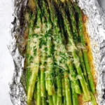 A close-up of delicious Parmesan grilled asparagus topped with melted cheese and fresh herbs, served in aluminum foil.