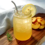 A mason jar filled with peach green tea lemonade, garnished with a lemon slice and metal straw, sits on a wooden board with fresh peach slices.