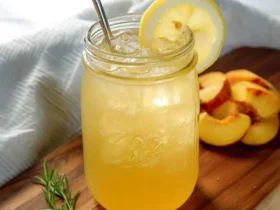 A mason jar filled with peach green tea lemonade, garnished with a lemon slice and metal straw, sits on a wooden board with fresh peach slices.
