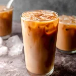 Close-up of a tall glass of creamy protein iced coffee with ice, featuring layered coffee and milk, on a textured grey surface.