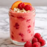 A vibrant Raspberry Peach Smoothie in a glass jar, topped with fresh raspberries, peach slices, and oats on a marble surface.