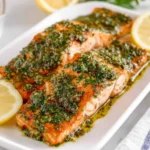 Two perfectly cooked salmon fillets are generously topped with vibrant green chimichurri sauce, served with fresh lemon slices on a white plate.