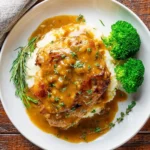 A perfectly cooked Skillet Chicken With Gravy served over creamy mashed potatoes, with fresh broccoli and herbs on a rustic wooden table.