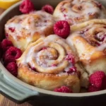 Sourdough Raspberry Lemon Rolls, glazed with white icing and fresh raspberries, in a baking dish with blurred lemons.