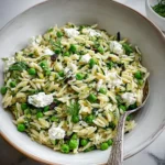 A large white bowl brimming with a vibrant Zucchini, Pea & Goat Cheese Orzo Salad, garnished with fresh dill and a silver spoon.