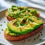 Two slices of avocado toast sandwich recipes topped with sliced avocado, chives, and red pepper flakes on a white plate.