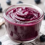 A glass jar filled with smooth, vibrant purple blueberry curd, surrounded by fresh blueberries.