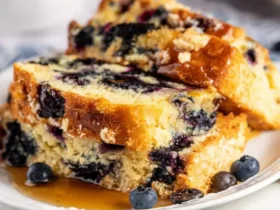 Stacked slices of Blueberry Pancake Bread drizzled with maple syrup on a white plate with fresh blueberries.