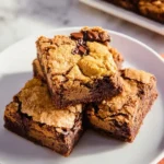 Three stacked brookie bars on a white plate, showing the chocolate chip cookie top and fudgy brownie layers.