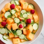 Fresh Cantaloupe Salad with Honey Lemon dressing, cucumber slices, raspberries, and mint in a white bowl.