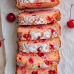 Sliced Cherry Almond Bread loaf dusted with powdered sugar on parchment paper with fresh cherries scattered around.