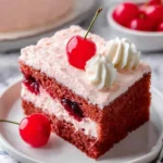 A slice of layered cherry cake with pink frosting, topped with a fresh cherry and whipped cream, served on a white plate.