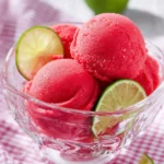 Scoops of vibrant Cherry Limeade Sorbet served in a glass bowl with fresh lime slices on a checkered pink tablecloth.