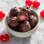 A bowl of Cherry Mash Truffles with one bitten to reveal the pink cherry center, surrounded by bright red cherries on marble.