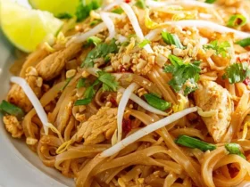 A plate of Chicken Pad Thai with rice noodles, bean sprouts, crushed peanuts, cilantro, and lime wedges.