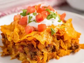 A slice of Doritos taco casserole topped with sour cream, diced tomatoes, and cilantro on a white plate.