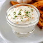 Creamy garlic aioli sauce in a glass bowl garnished with chives and paprika, served with toasted bread.
