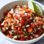 Fresh Pico de Gallo salsa with diced tomatoes, onions, and cilantro in a white bowl with lime wedges.
