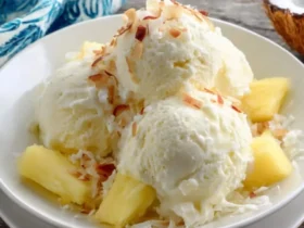Three scoops of creamy Pina Colada Ice Cream topped with toasted coconut flakes and fresh pineapple chunks in a white bowl.