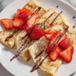 Three folded strawberry crepes topped with fresh sliced strawberries, chocolate drizzle, and powdered sugar on a white plate.