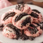 Stack of pink Strawberry Oreo Cheesecake Cookies topped with crushed Oreos and cream filling on a white plate.